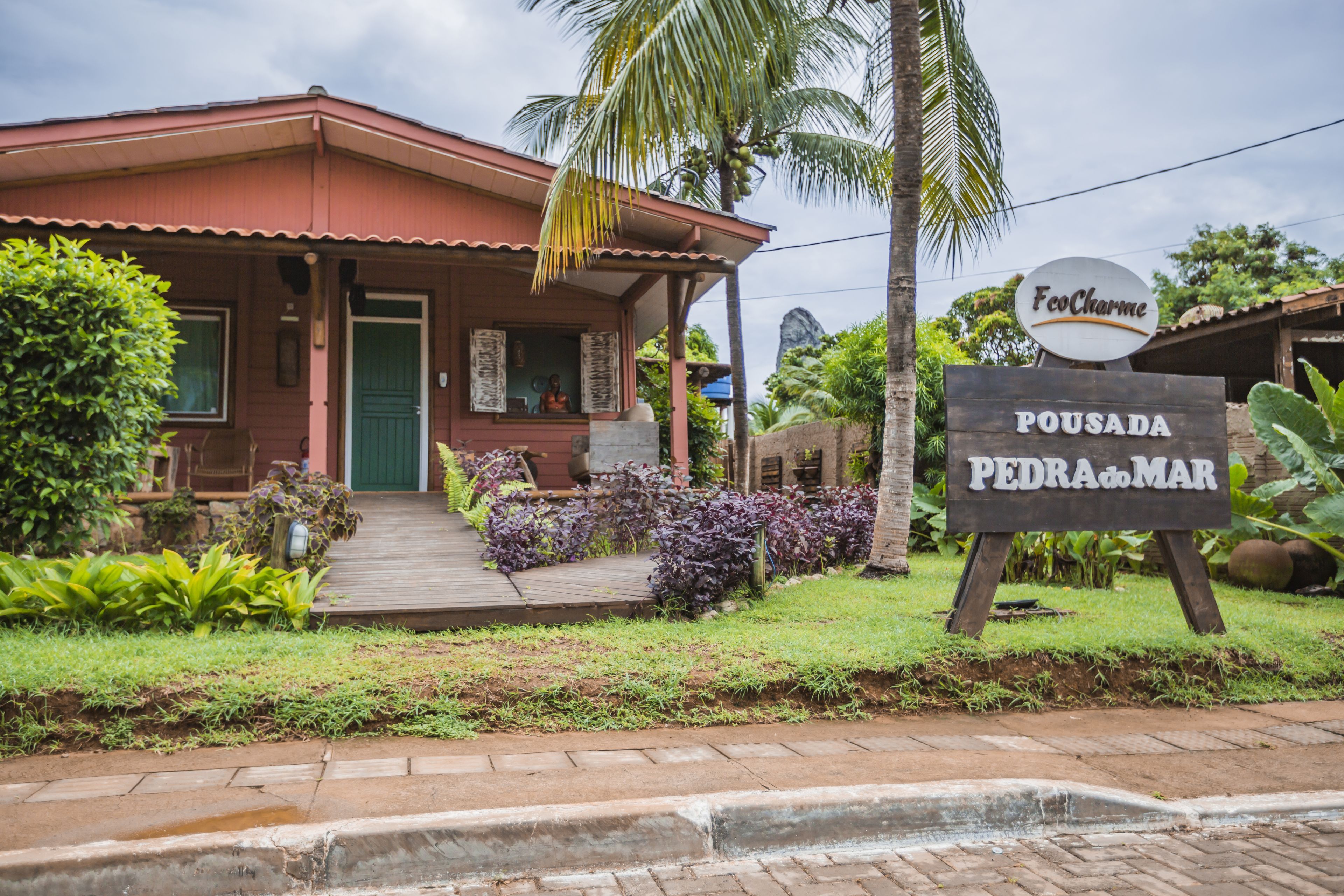 A house with a porch, a sign reading "EcoCharme Pousada Pedra do Mar", and lush greenery around.
