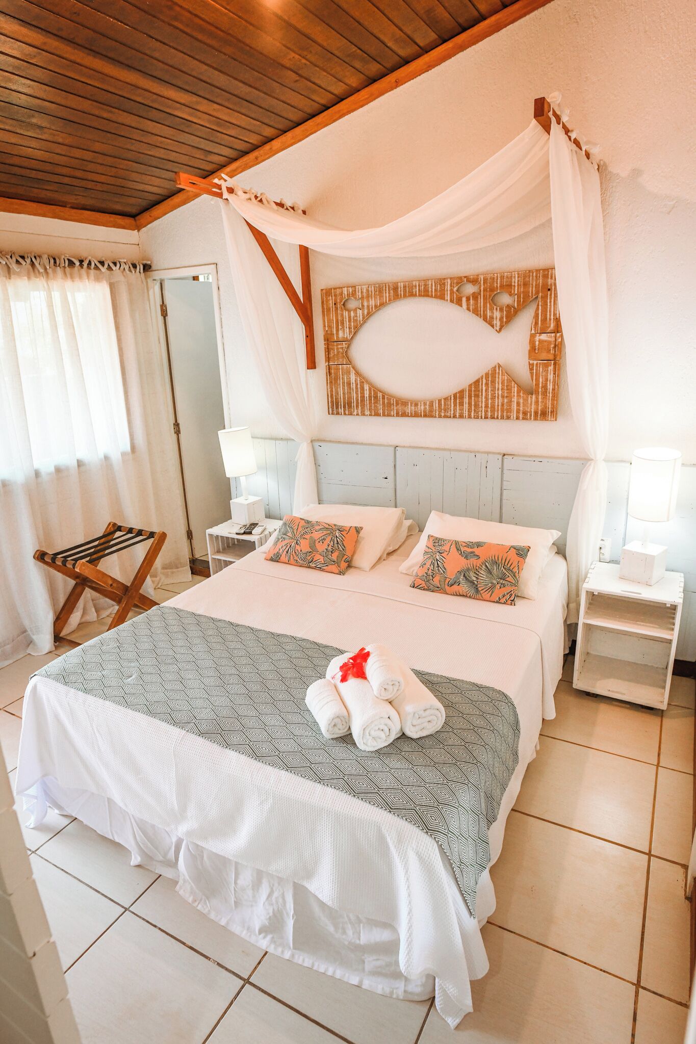 A bedroom with a bed, white curtains, a wooden ceiling, a wall art of a fish, and a bedside table with a lamp.