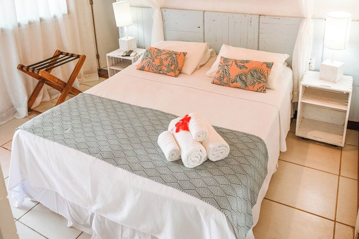 A bedroom with a bed, white curtains, a wooden ceiling, a wall art of a fish, and a bedside table with a lamp.