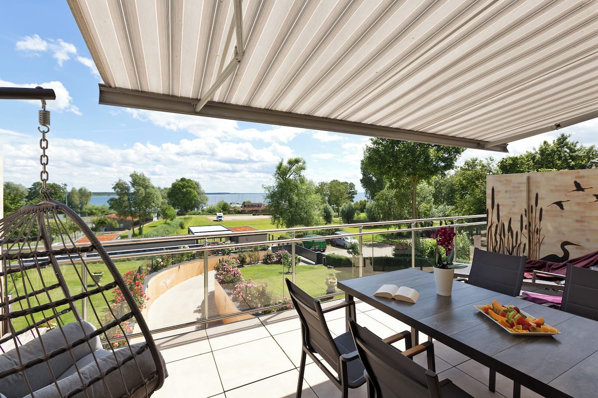 Ein Balkon mit Blick auf einen Garten und einen See, ein Tisch mit Stühlen und einem Hängesessel.
