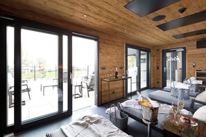 Un intérieur moderne avec un plafond en bois, de grandes portes-fenêtres coulissantes donnant sur un patio extérieur, un coin salon avec une table en verre et un bar avec une bouteille de champagne.