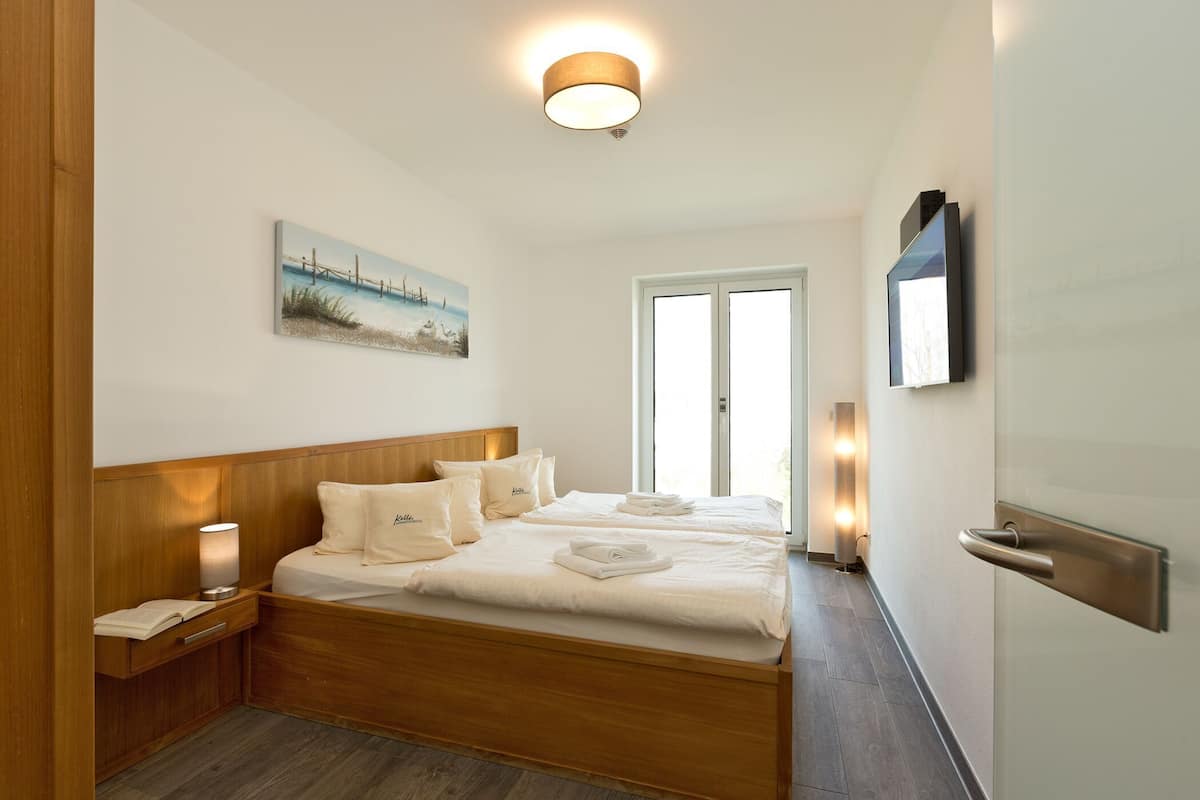 Ein Hotelzimmer mit einem hölzernen Kopfende, einem Bett mit ordentlich bezogenen Leinen, einem Nachttisch mit Lampe, einem Bild an der Wand und einem an der Wand befestigten Fernseher.