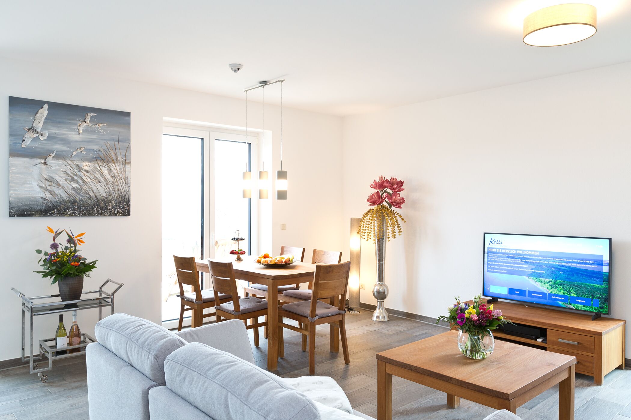 A modern living room with a sofa, dining table, chairs, a TV, and a vase of flowers.