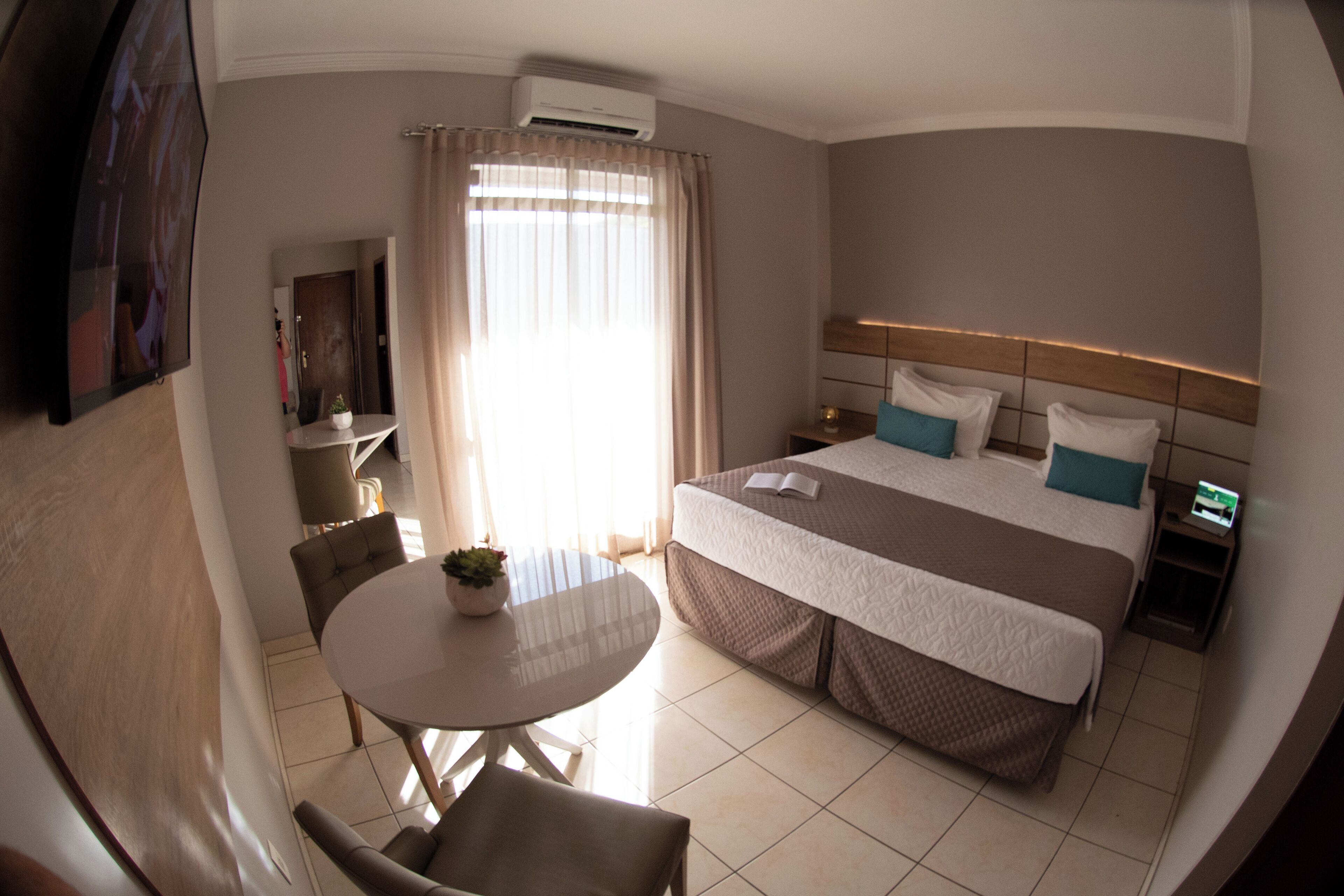 Habitación de hotel con cama, mesitas de noche, televisor, una mesa de comedor con sillas y una ventana con cortinas.