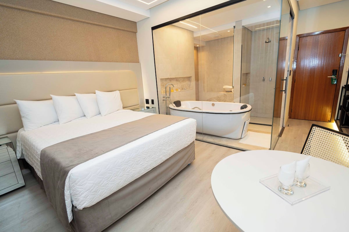 A modern hotel room with a large bed, a separate bathroom with a bathtub, and a round dining table.