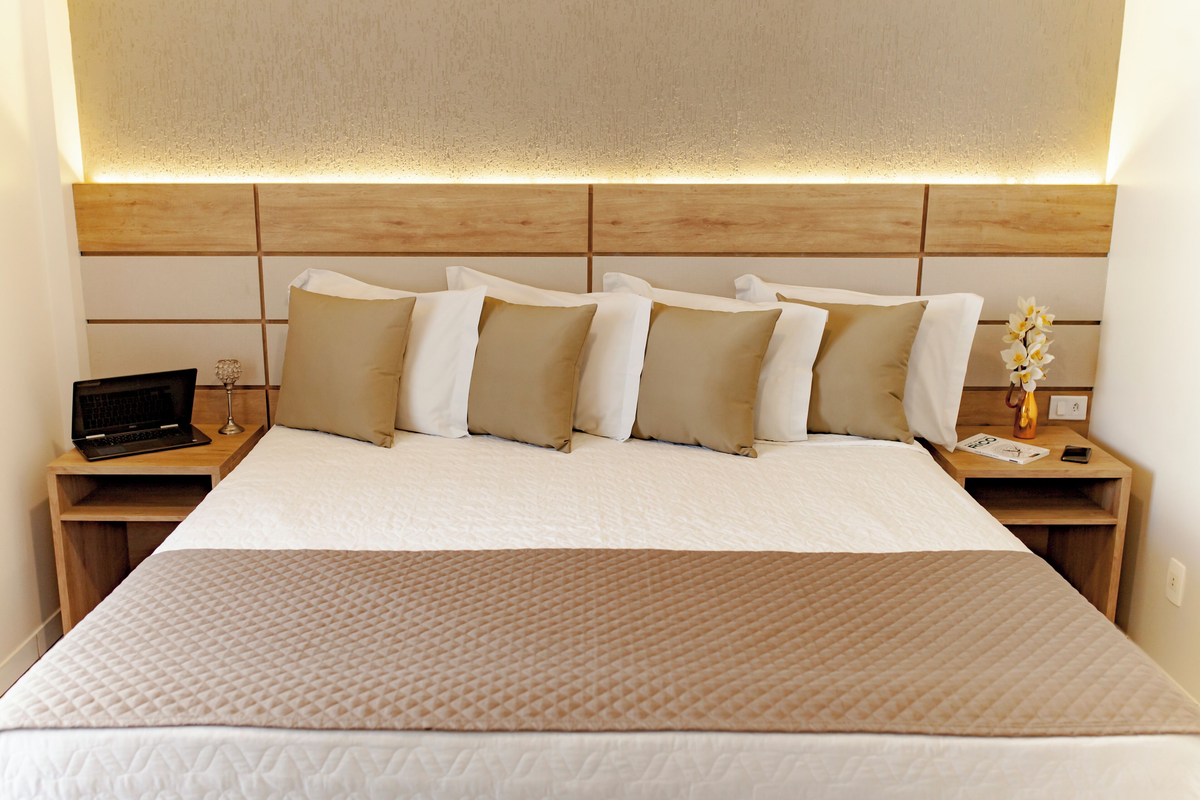 A neatly made bed with a headboard, a bedside table with a laptop, and a small vase with flowers.