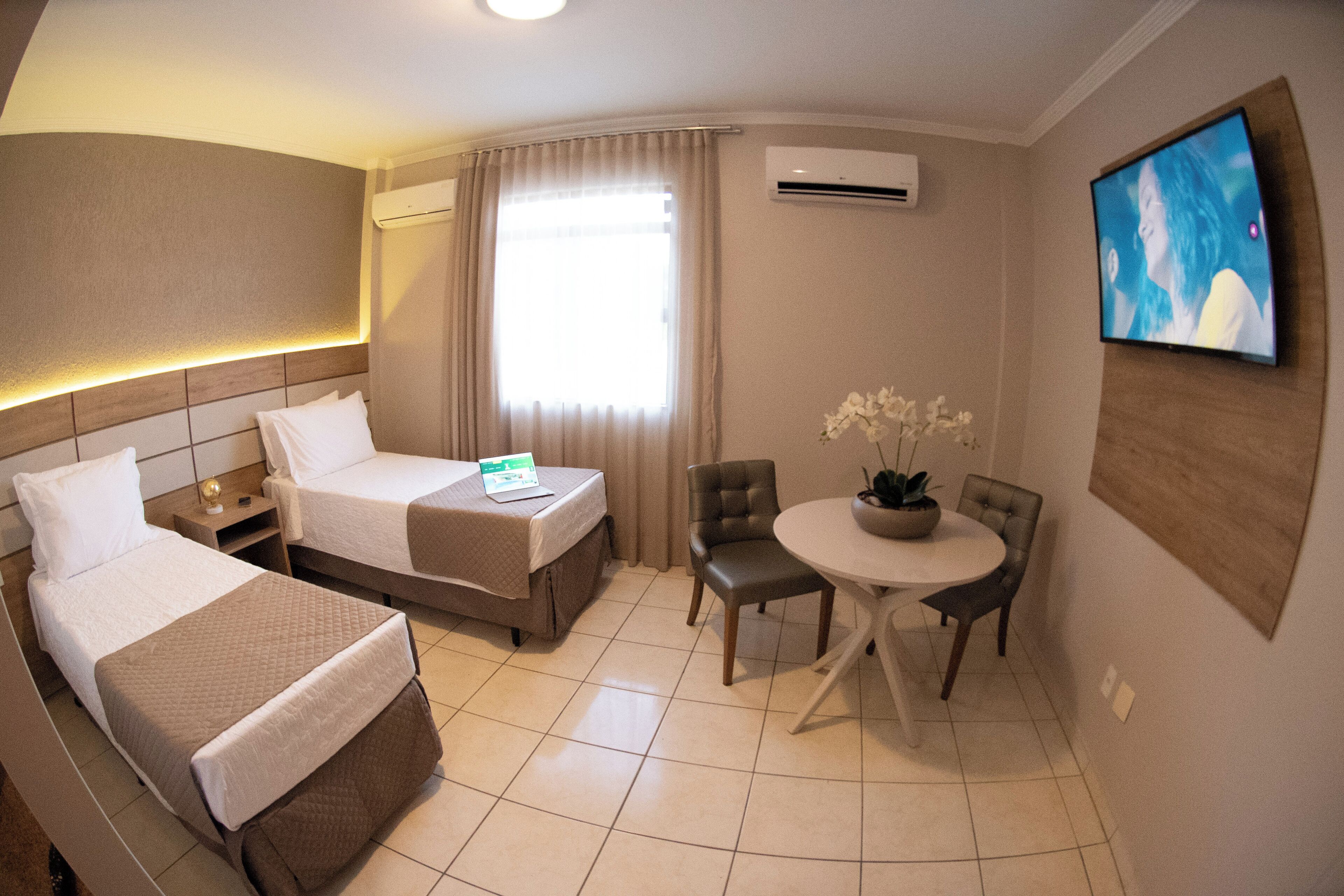 A hotel room with two beds, a dining table with chairs, a TV mounted on the wall, and an air conditioning unit.