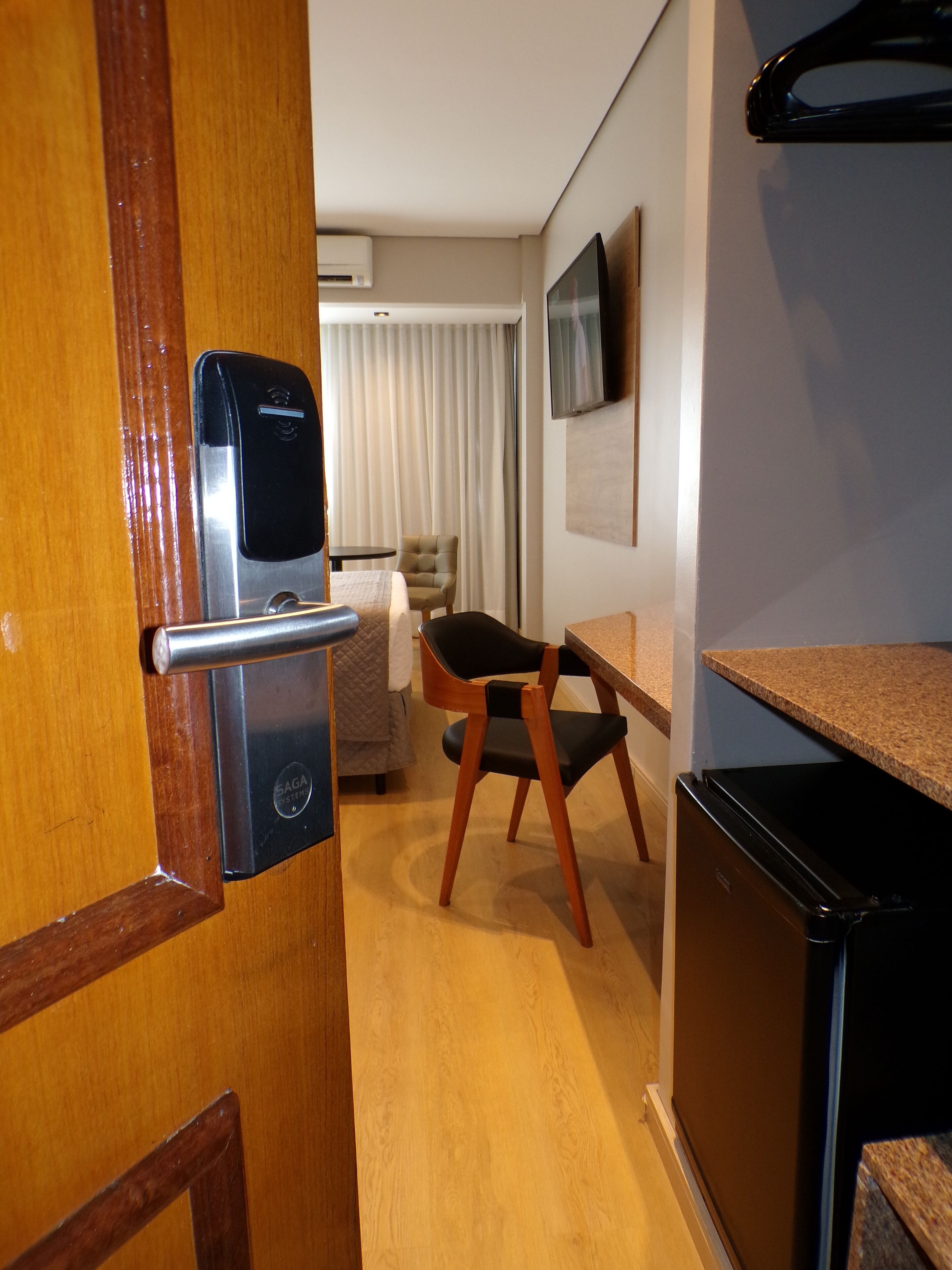 A compact hotel room with a wooden door, a minibar, a small refrigerator, a bed with a white duvet, a chair, and a television mounted on the wall.