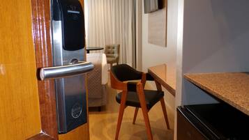 A compact hotel room with a wooden door, a minibar, a small refrigerator, a bed with a white duvet, a chair, and a television mounted on the wall.