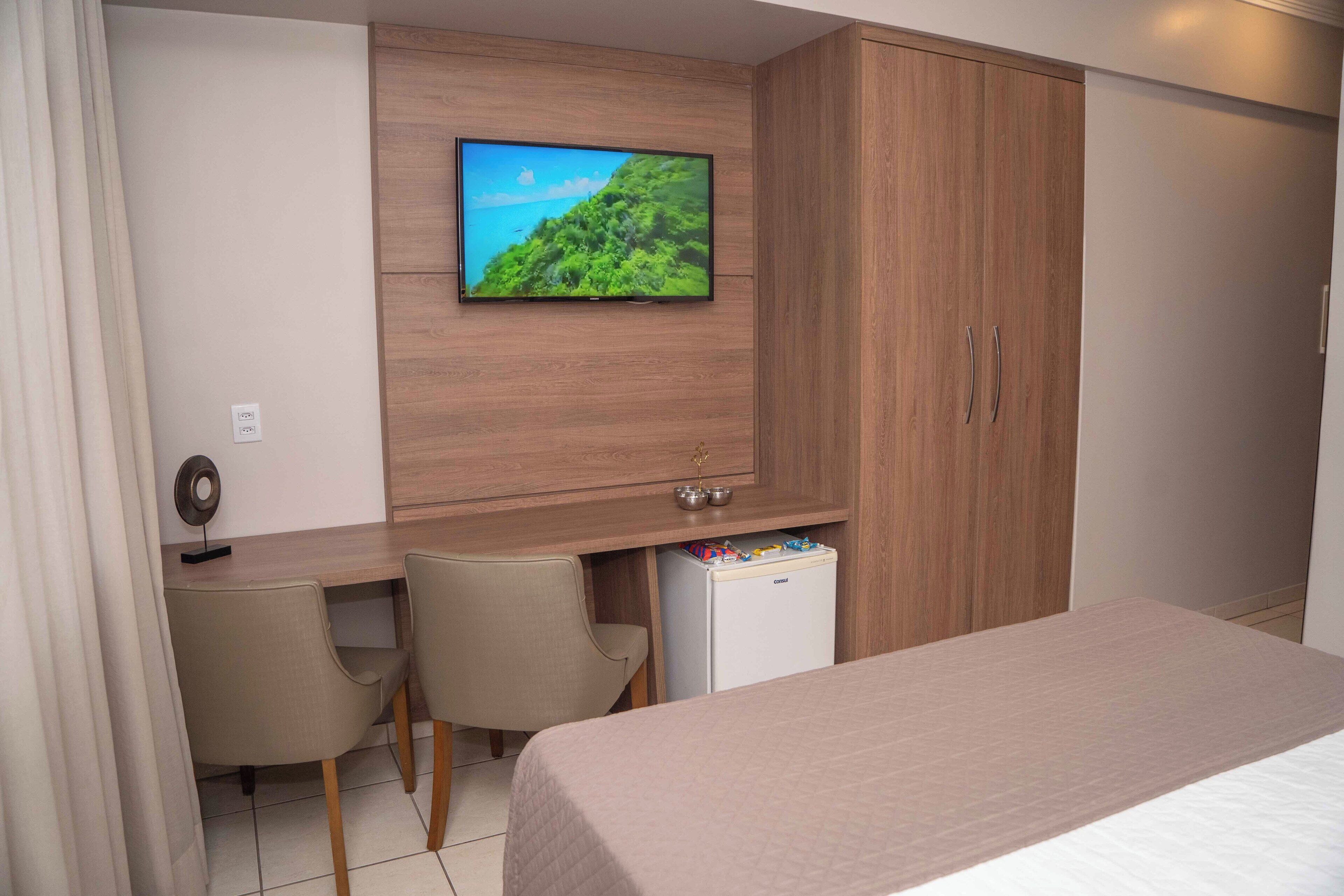 Ein Hotelzimmer mit einem Bett, einem Schreibtisch mit zwei Stühlen, einem an der Wand befestigten Fernseher und einer Minibar.