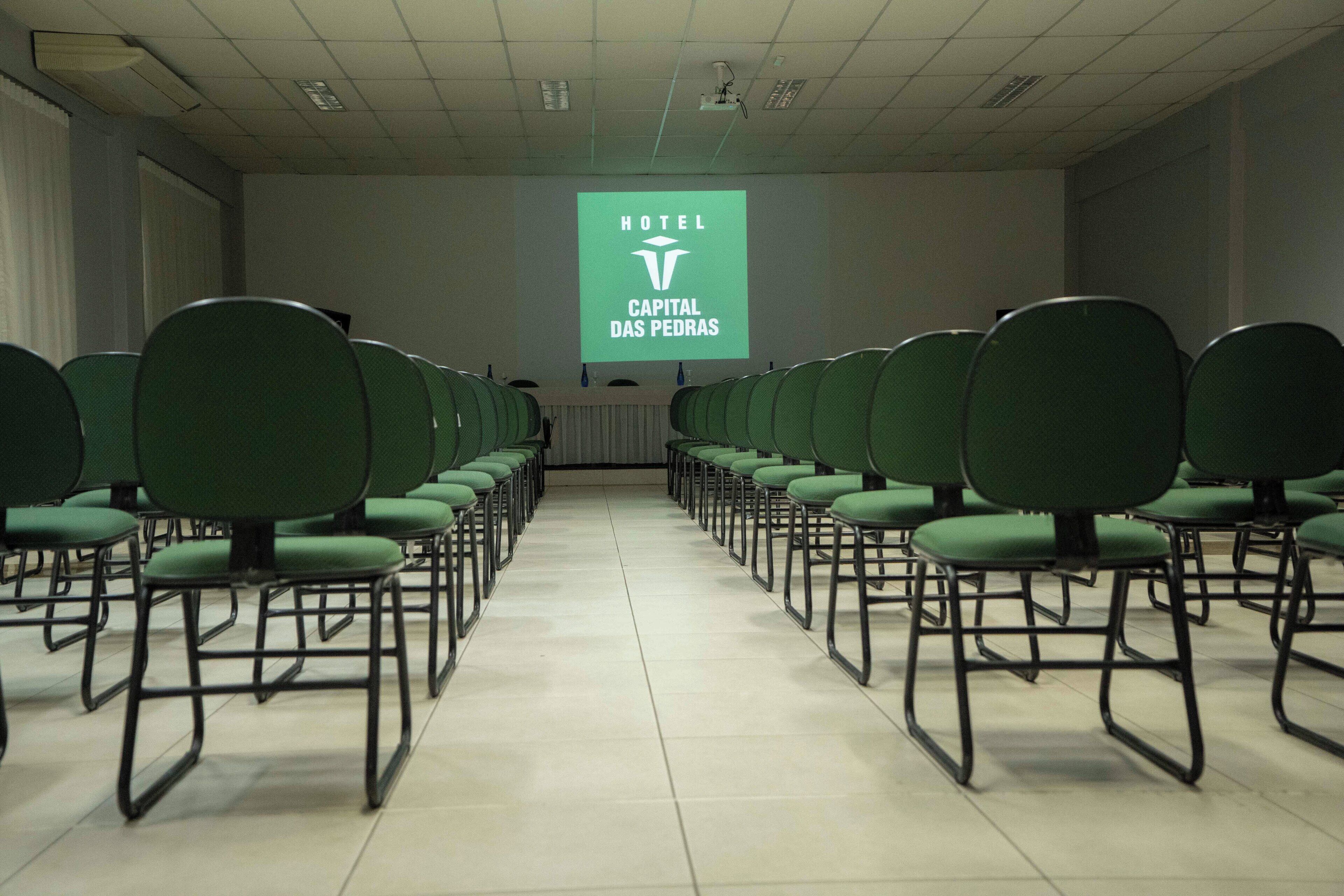 A conference room with rows of green chairs and a projection screen displaying "HOTEL CAPITAL DAS PEDRAS".