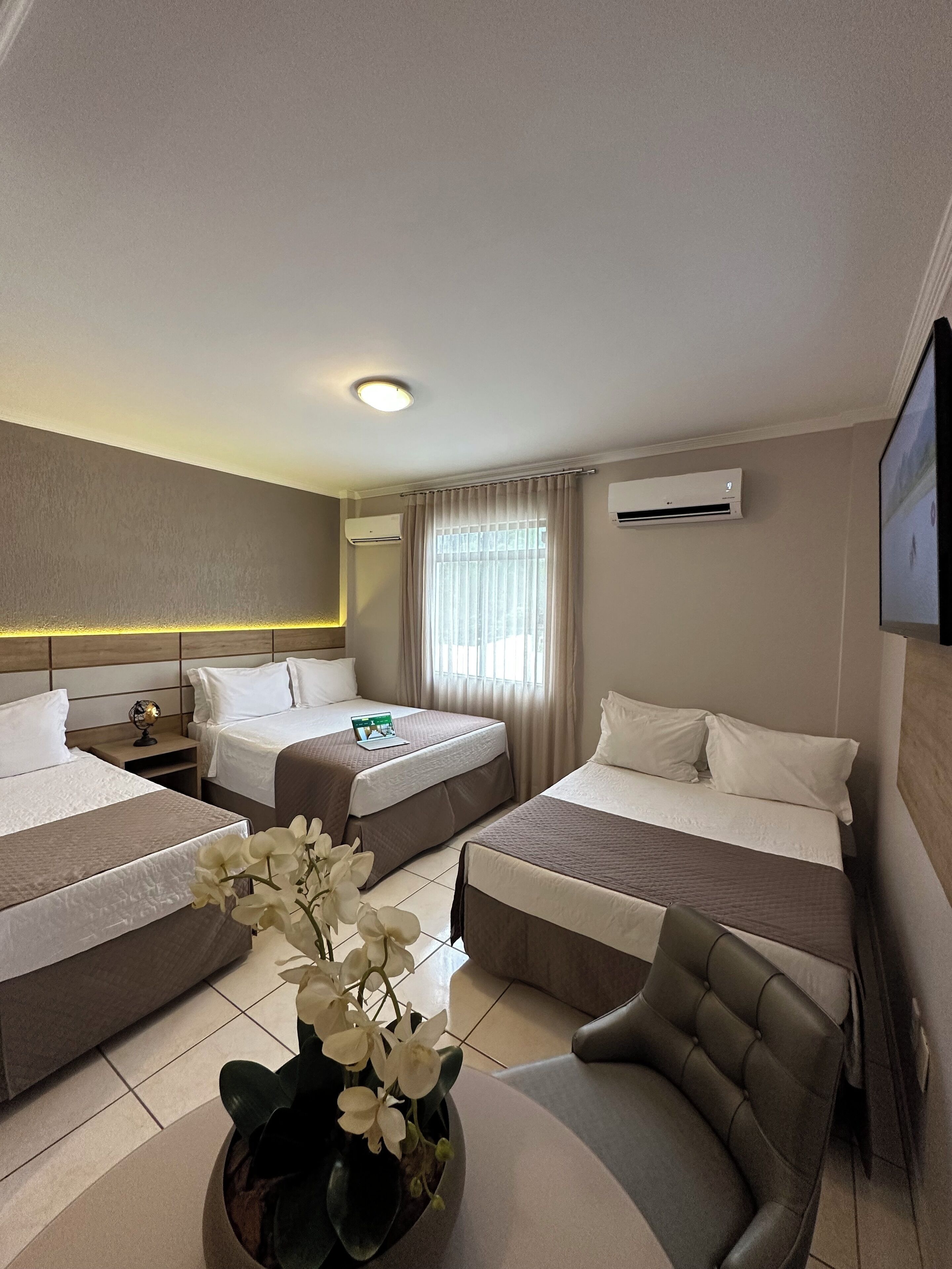A hotel room with two beds, a desk, a chair, a potted orchid, and an air conditioning unit.