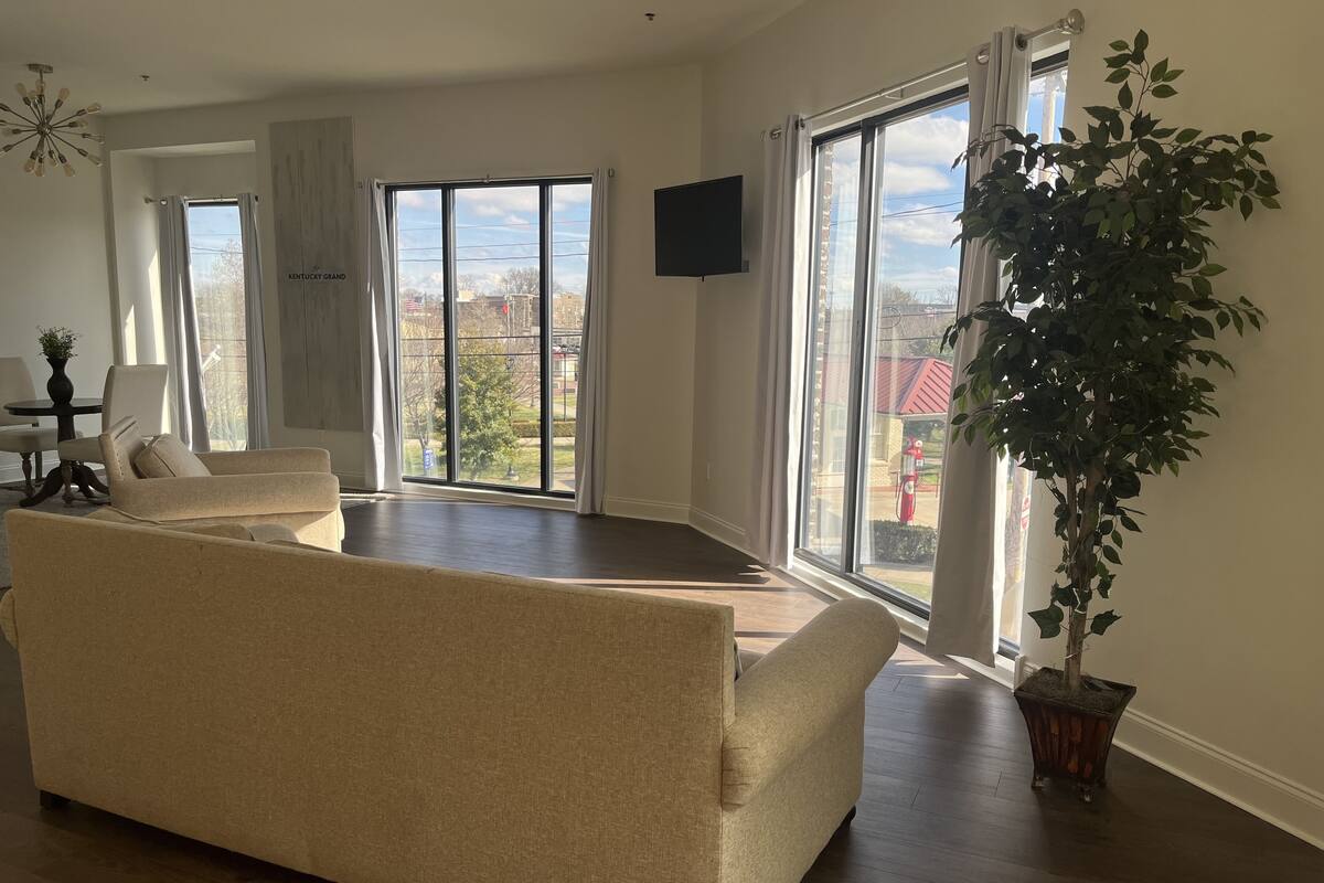 Ein geräumiges Wohnzimmer mit einem beigen Sofa, einem Esstisch mit Stühlen, einem großen Fenster mit Blick auf das Kentucky Grand und einer Zimmerpflanze.