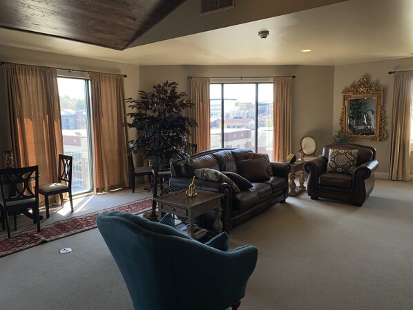 A spacious living room with a leather sofa, a blue armchair, a large potted plant, and a dining area with a red carpet. - Kentucky Grand Hotel & Spa (Bowling Green)