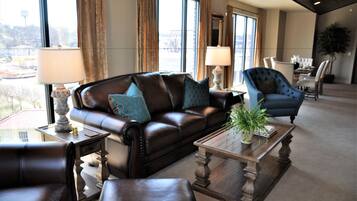 A modern living room with a leather sofa, armchairs, and a coffee table. There are two lamps on side tables, and a large window provides a view of the city.