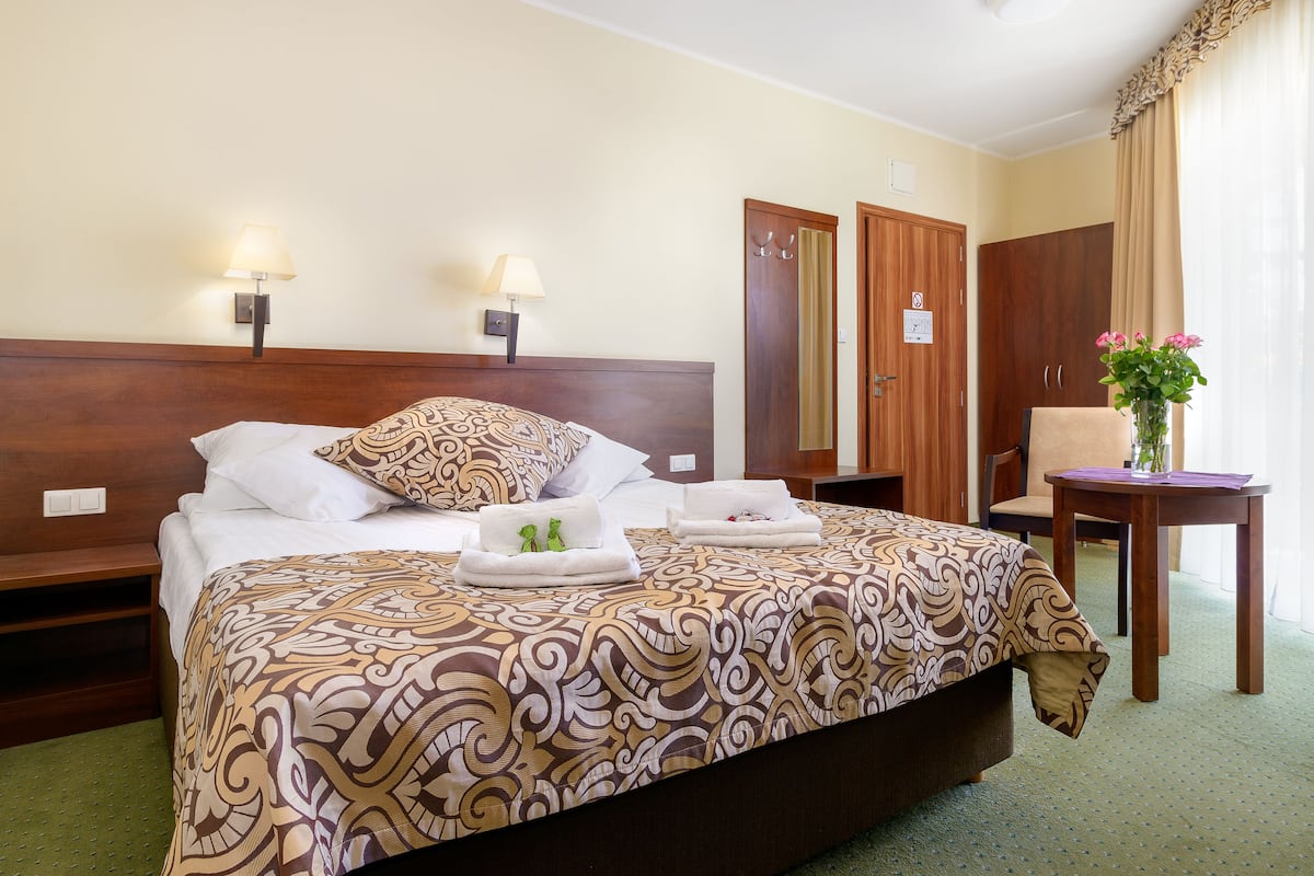 Quarto de hotel com uma cama grande, duas luminárias de cabeceira, um guarda-roupa de madeira, uma mesinha com um vaso de flores e um tapete verde.