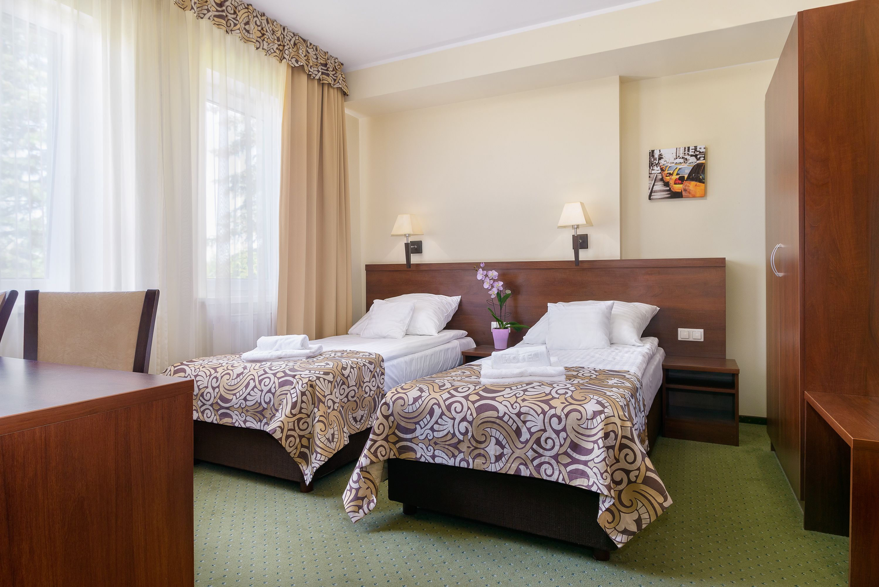 A hotel room with two beds, a desk, and a chair. There are two bedside tables with lamps, a potted plant, and a painting on the wall.