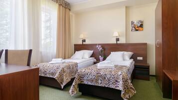 A hotel room with two beds, a desk, and a chair. There are two bedside tables with lamps, a potted plant, and a painting on the wall.