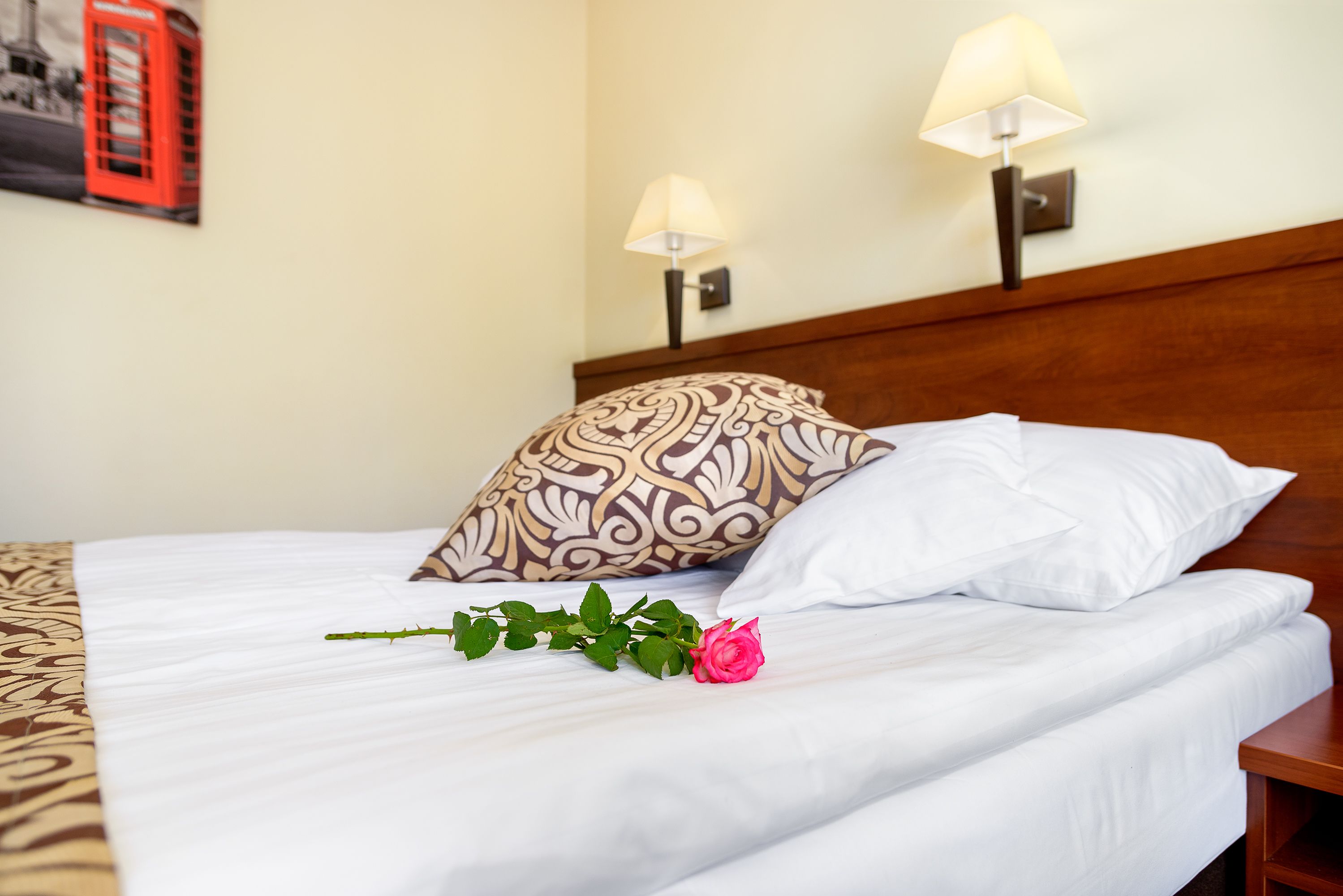 A neatly made bed with white linens, a patterned pillow, and a single rose.