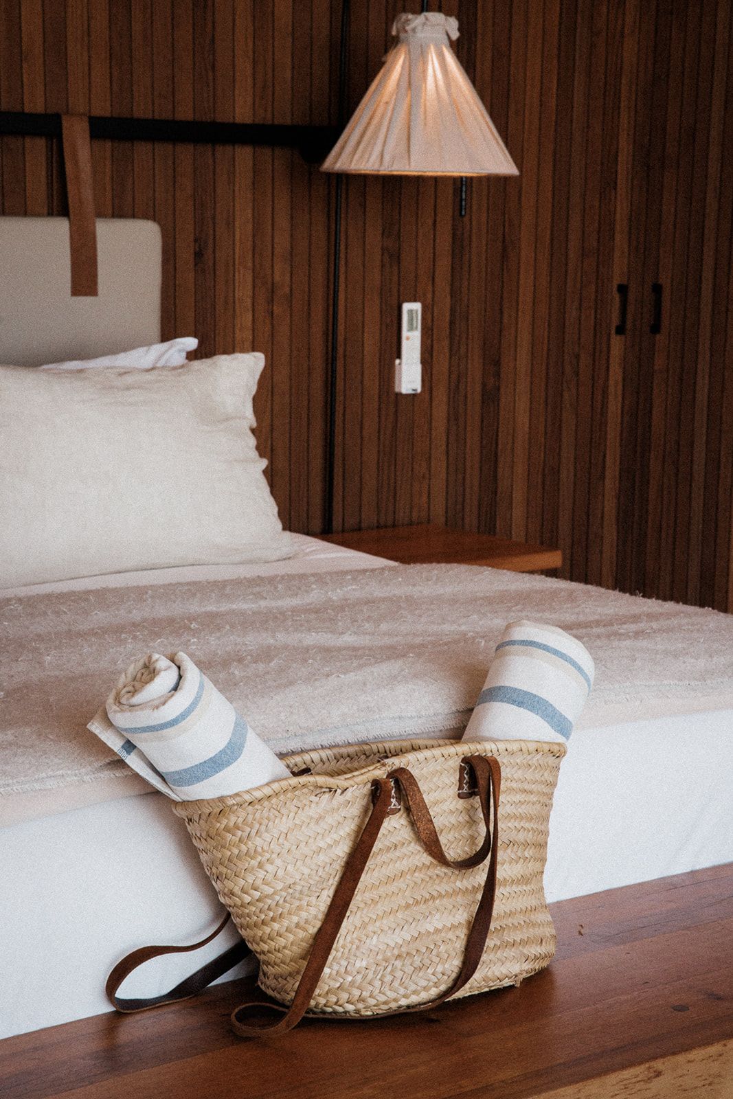 A bed with a white comforter, a wooden headboard, a woven basket with a brown strap, and a wall-mounted lamp.