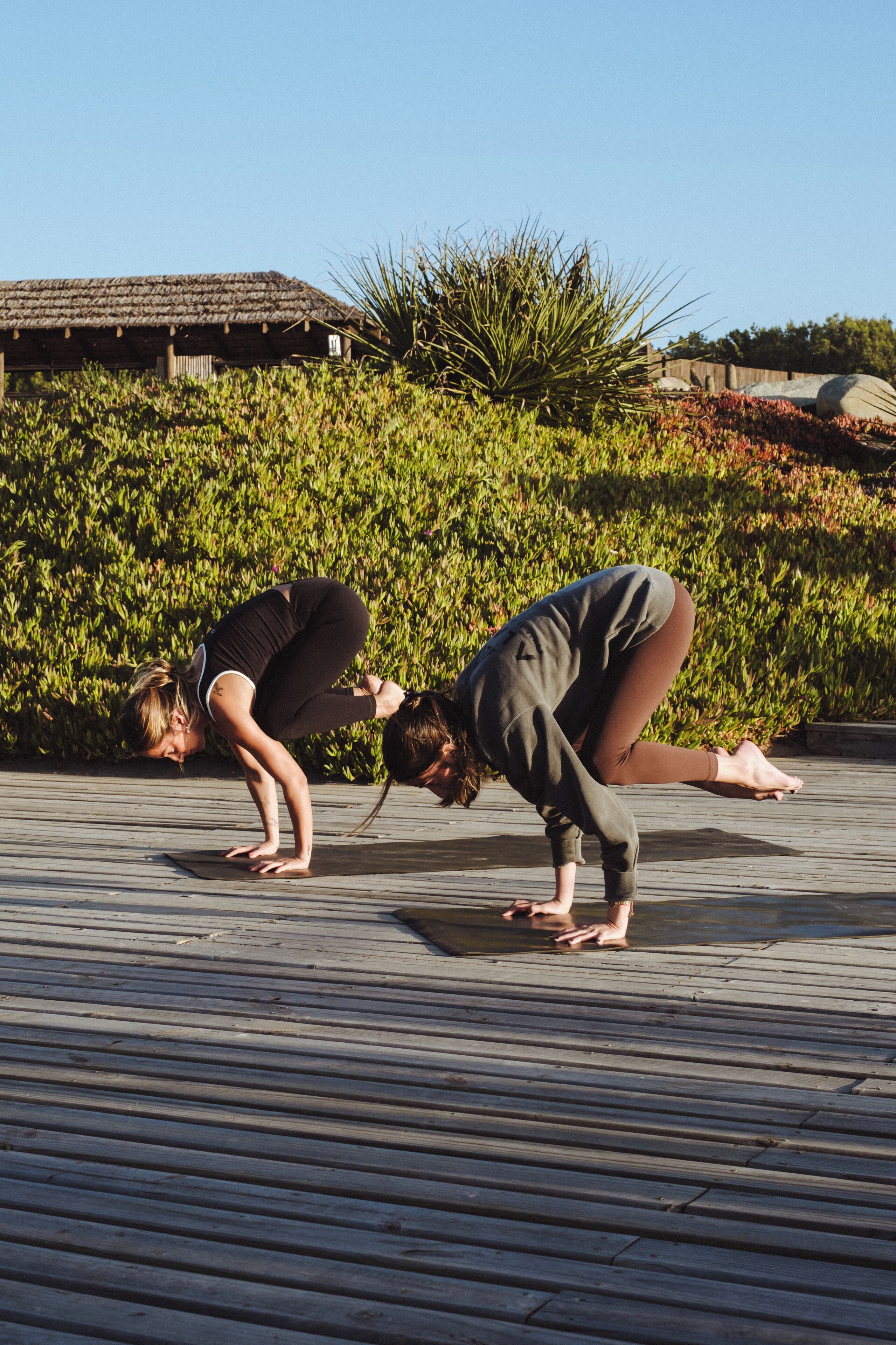 Dos personas realizando una postura de yoga sobre una terraza de madera, con un fondo de vegetación y un edificio.