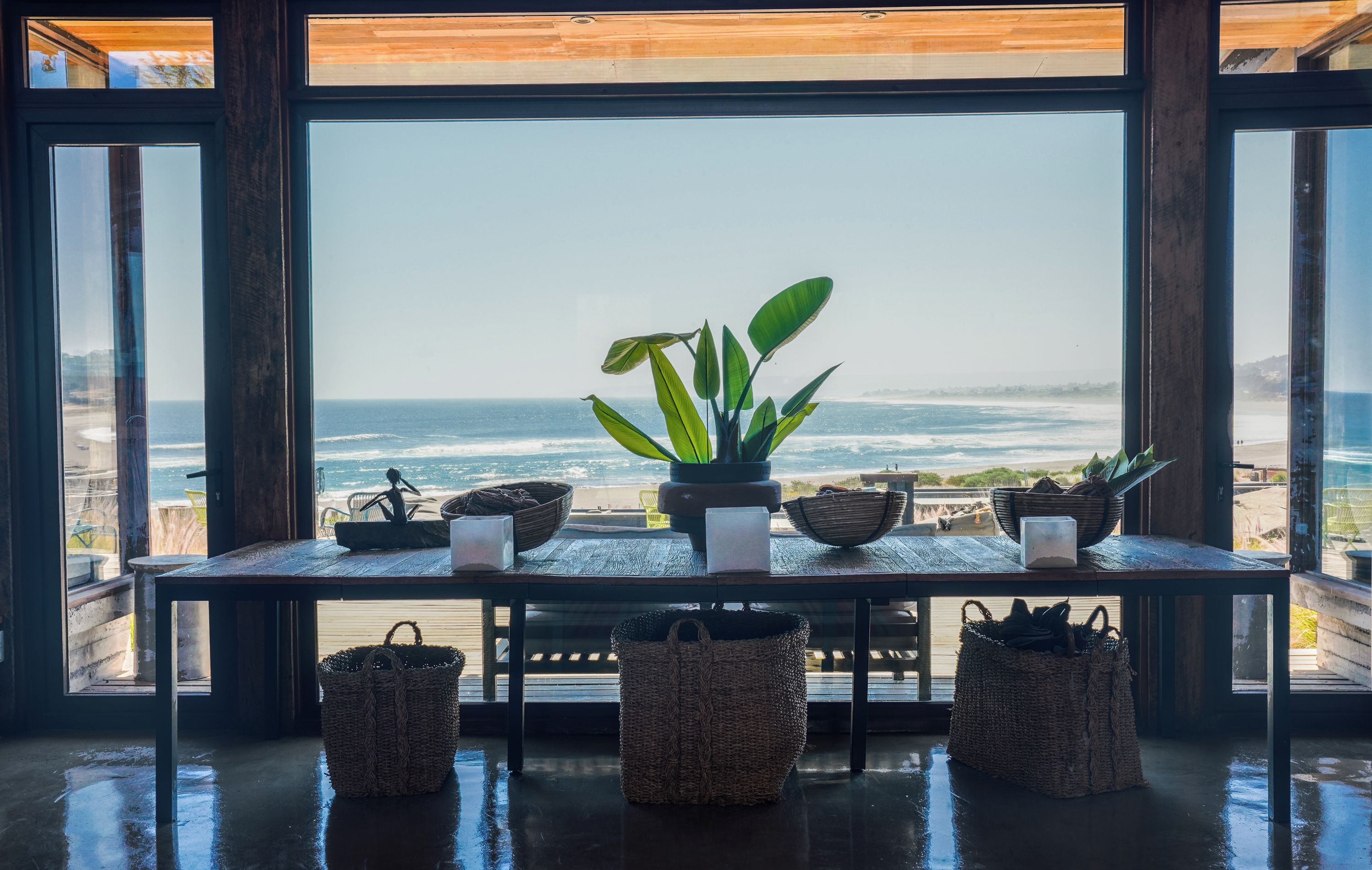 A room with a large window overlooking a beach, a wooden table with baskets and a plant, and a sculpture.