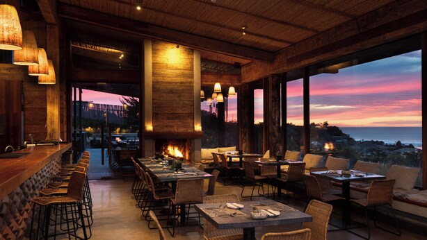 A restaurant with a long bar, multiple tables, and chairs, with a view of the ocean at sunset. - Hotel Alaia (Pichilemu)