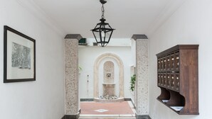 A hallway with a tiled floor, a framed picture on the left, a decorative light fixture, a stone fountain, and a wooden cabinet. - Habitat's Campo di Fiori Apartments (Rome)