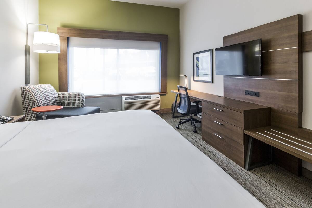 A hotel room with a large bed, a desk with a chair, a TV mounted on the wall, and a window with blinds.