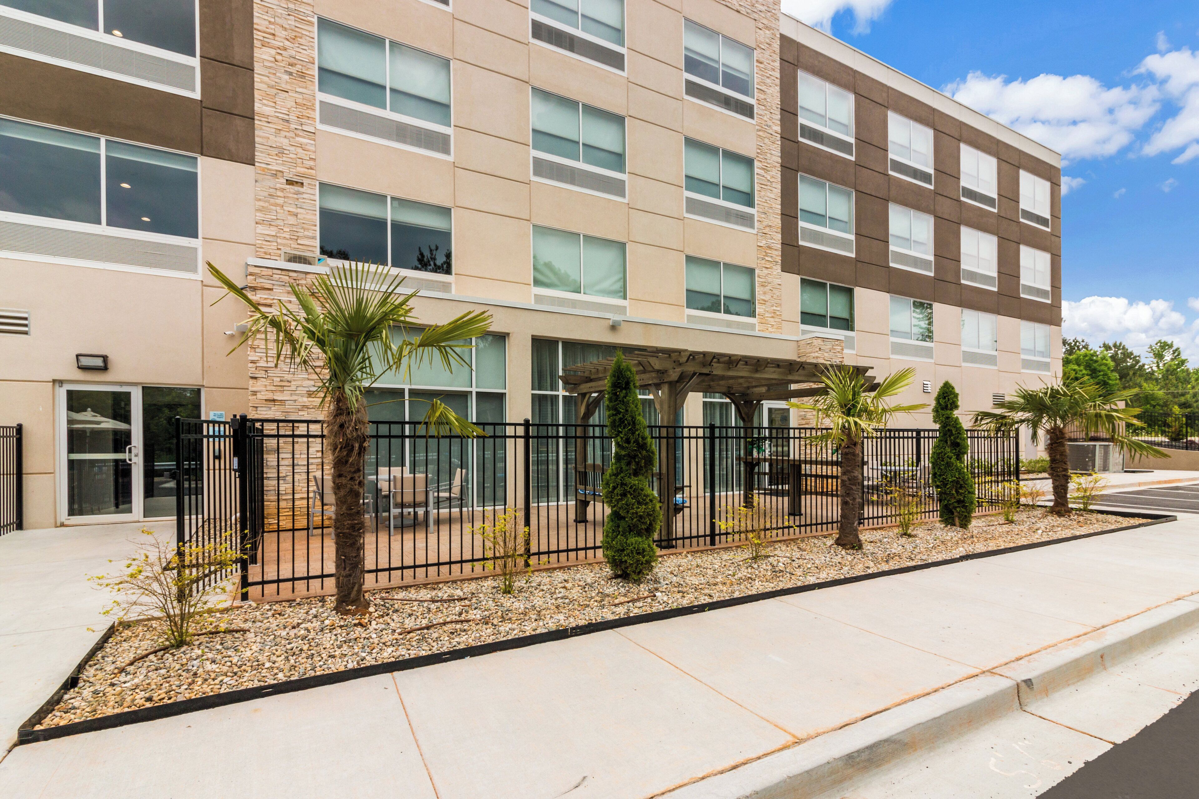 A multi-story building with a modern architectural design, featuring a mix of stone and glass facade, outdoor seating area with palm trees, and a gated entrance.