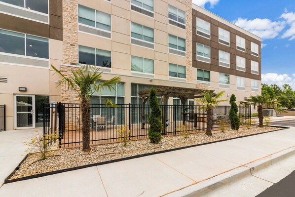 A multi-story building with a modern architectural design, featuring a mix of stone and glass facade, outdoor seating area with palm trees, and a gated entrance.