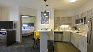 Quarto de hotel com cama, escrivaninha, cadeira, pia em um móvel compacto, micro-ondas, frigorífico e área de refeições com uma cadeira amarela.