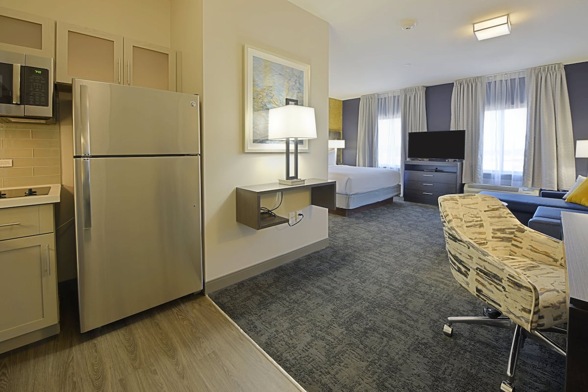 Hotelkamer met kitchenette, een bed, televisie, bureau met stoel en groot raam met gordijnen.