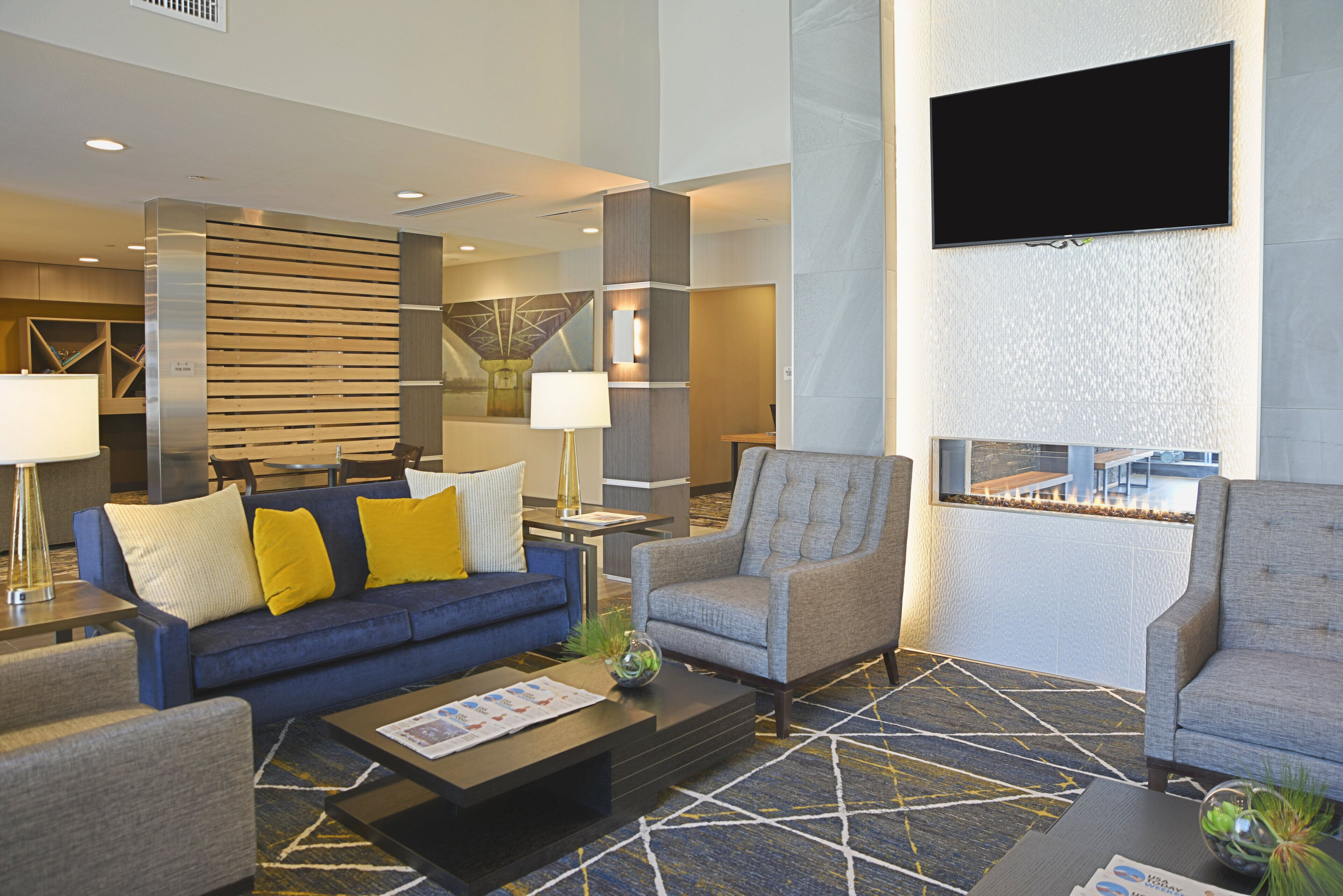 A modern lobby with a blue sofa, armchairs, a coffee table, and a flat-screen TV mounted on the wall.