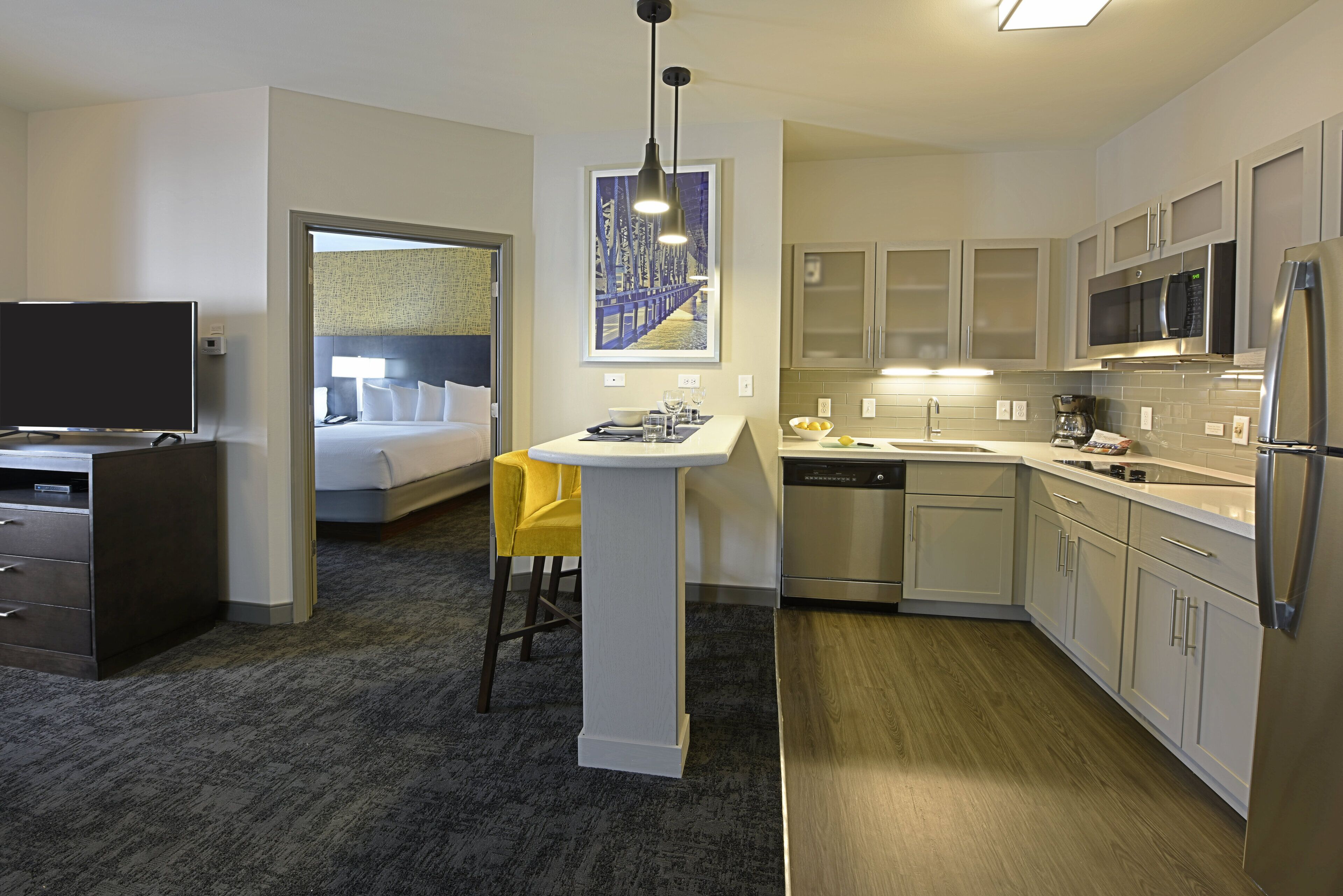 A hotel room with a bed, a desk, a chair, a small kitchenette, and a bathroom.