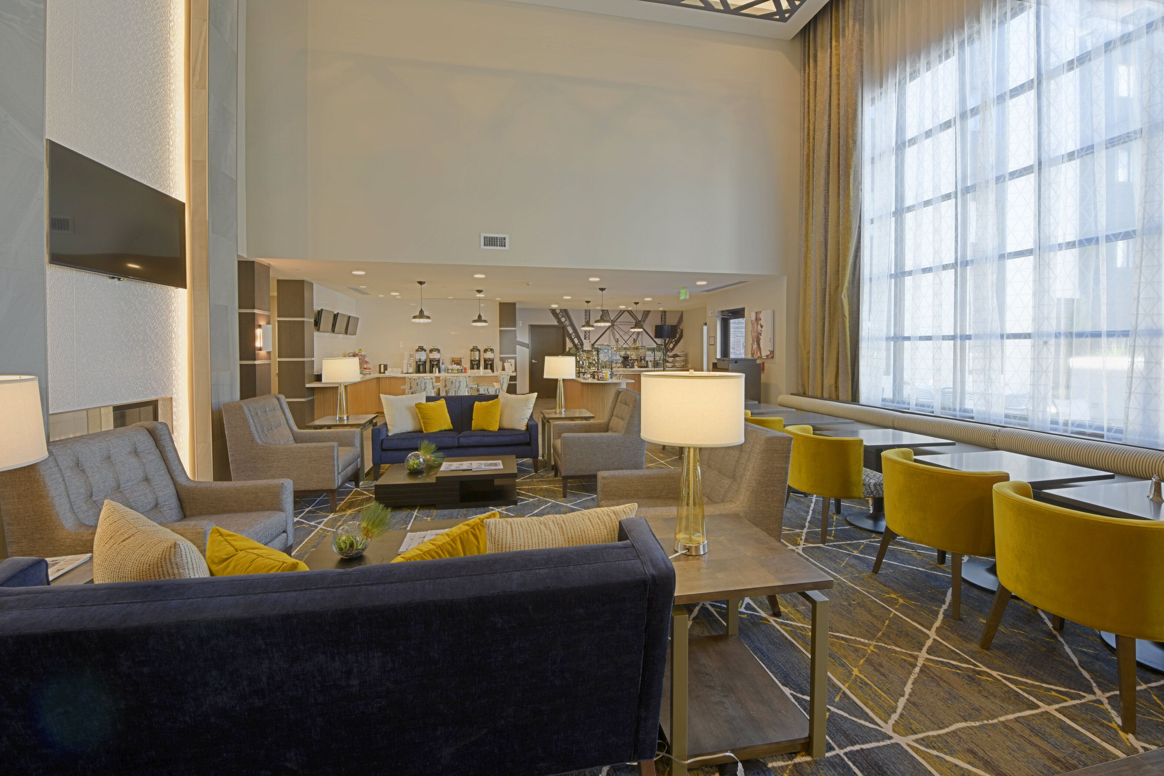 Un moderno vestíbulo de hotel con zona de estar, una cafetería y un amplio ventanal que proporciona luz natural.