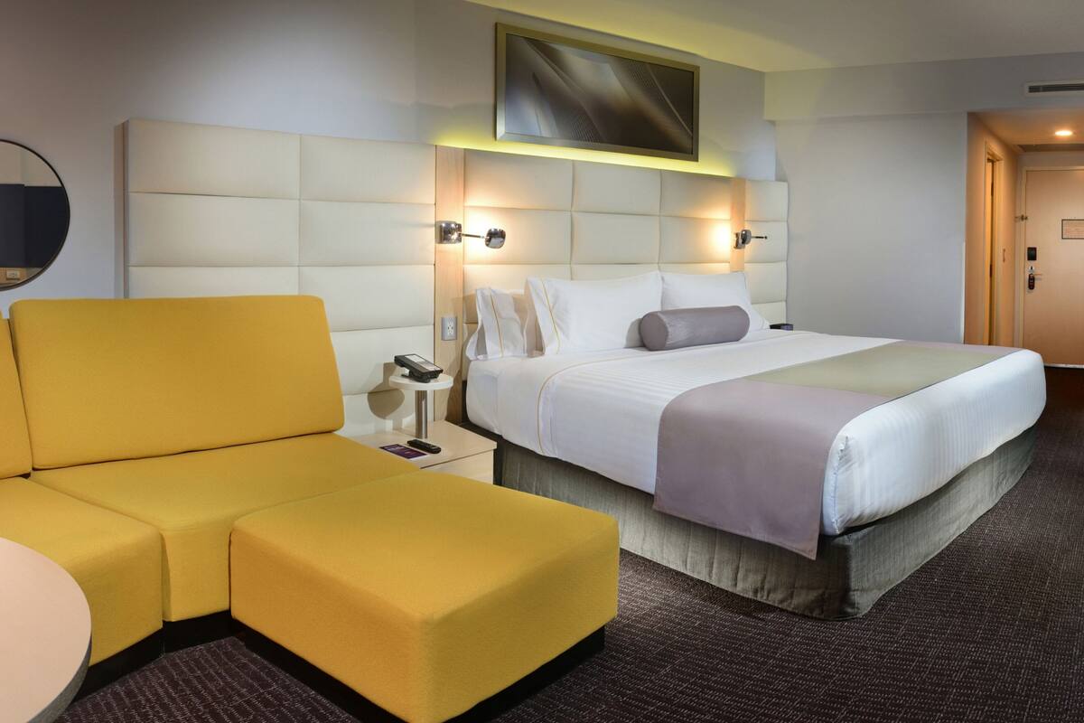 A hotel room with a large bed, a yellow sofa, a small round table, and a wall-mounted artwork.