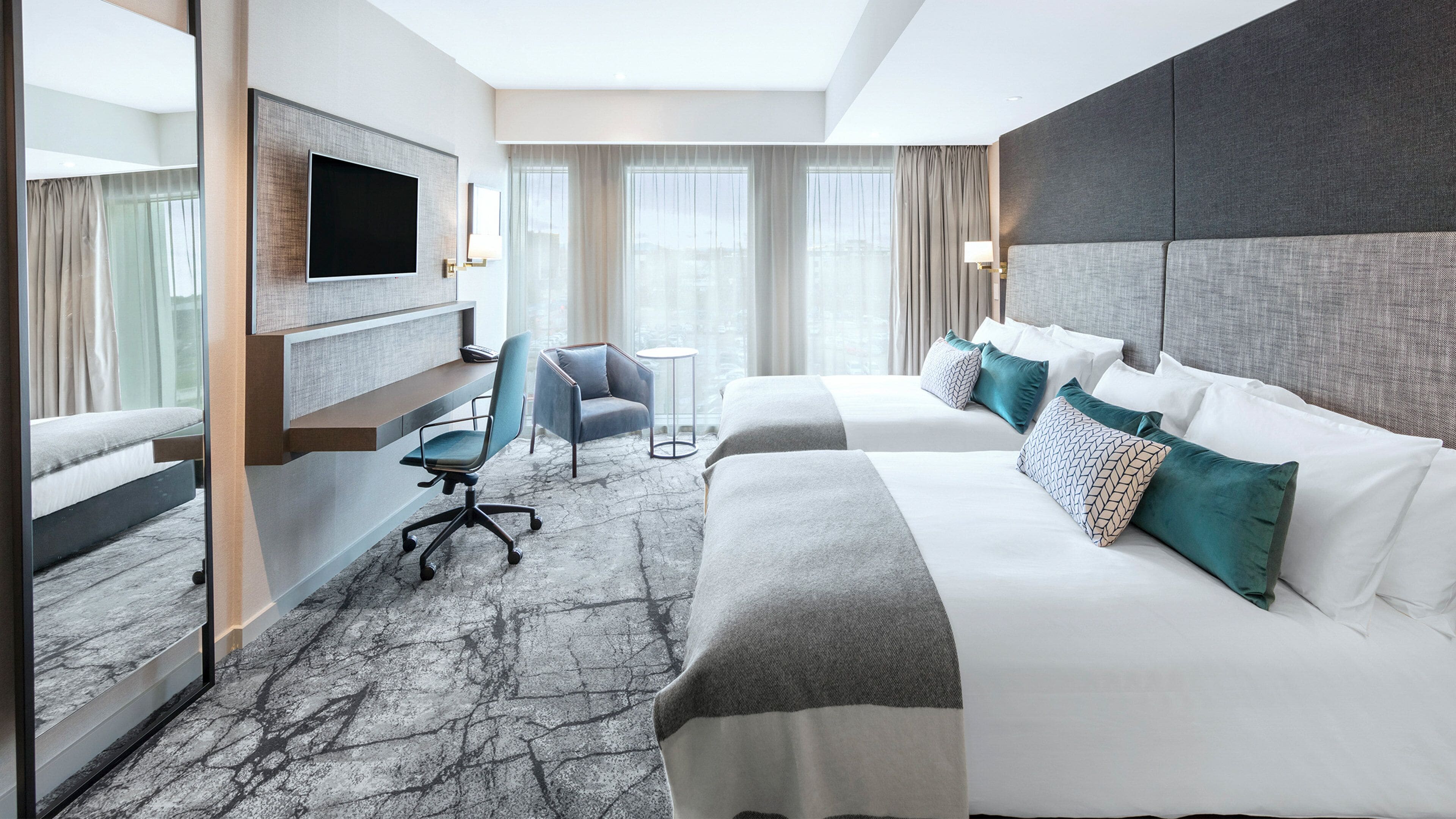 Une chambre d’hôtel avec un grand lit, un lit plus petit, un bureau avec une chaise, une télévision et une vue sur la ville grâce à de grandes fenêtres.