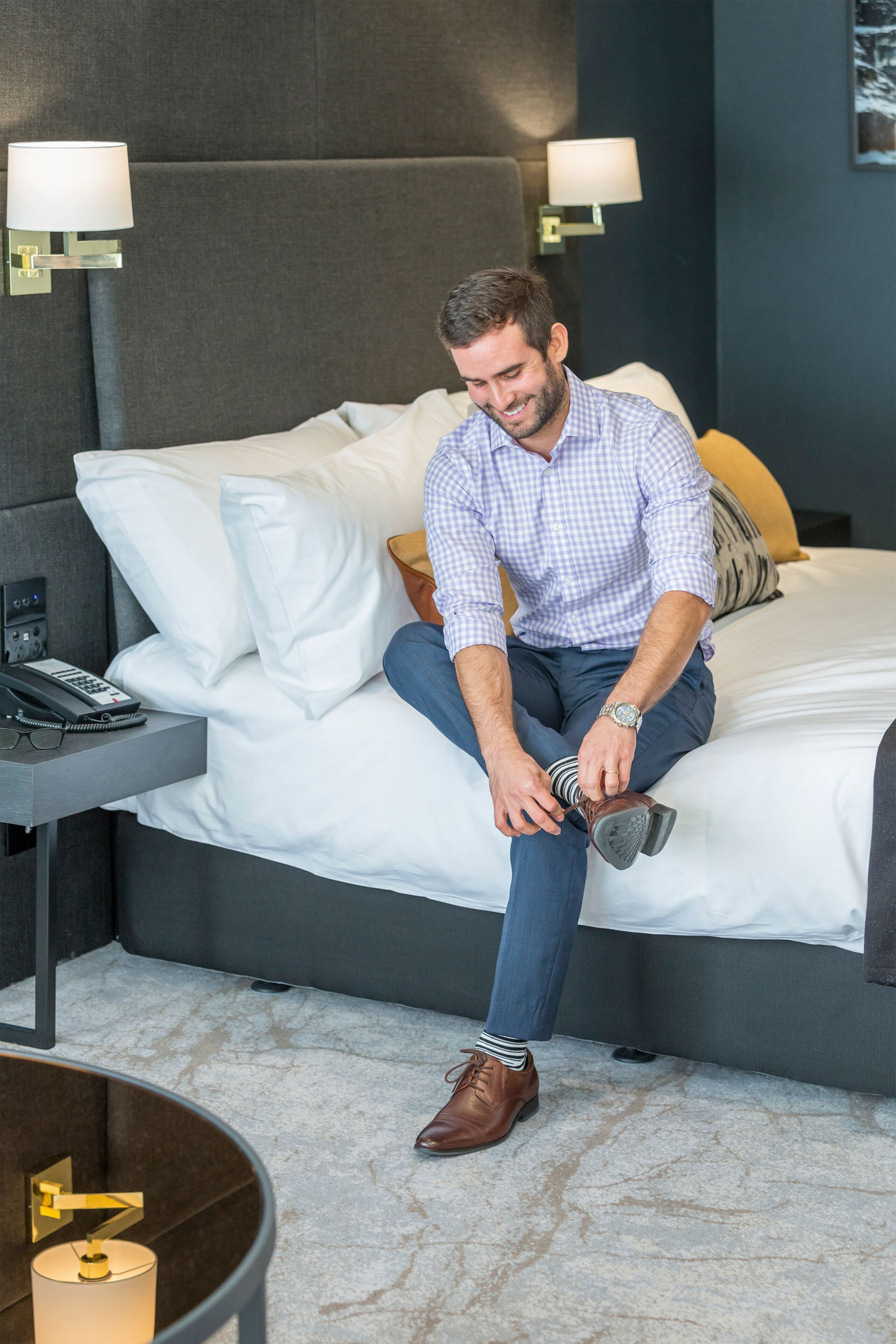 A man sitting on a bed in a hotel room, adjusting his shoe.