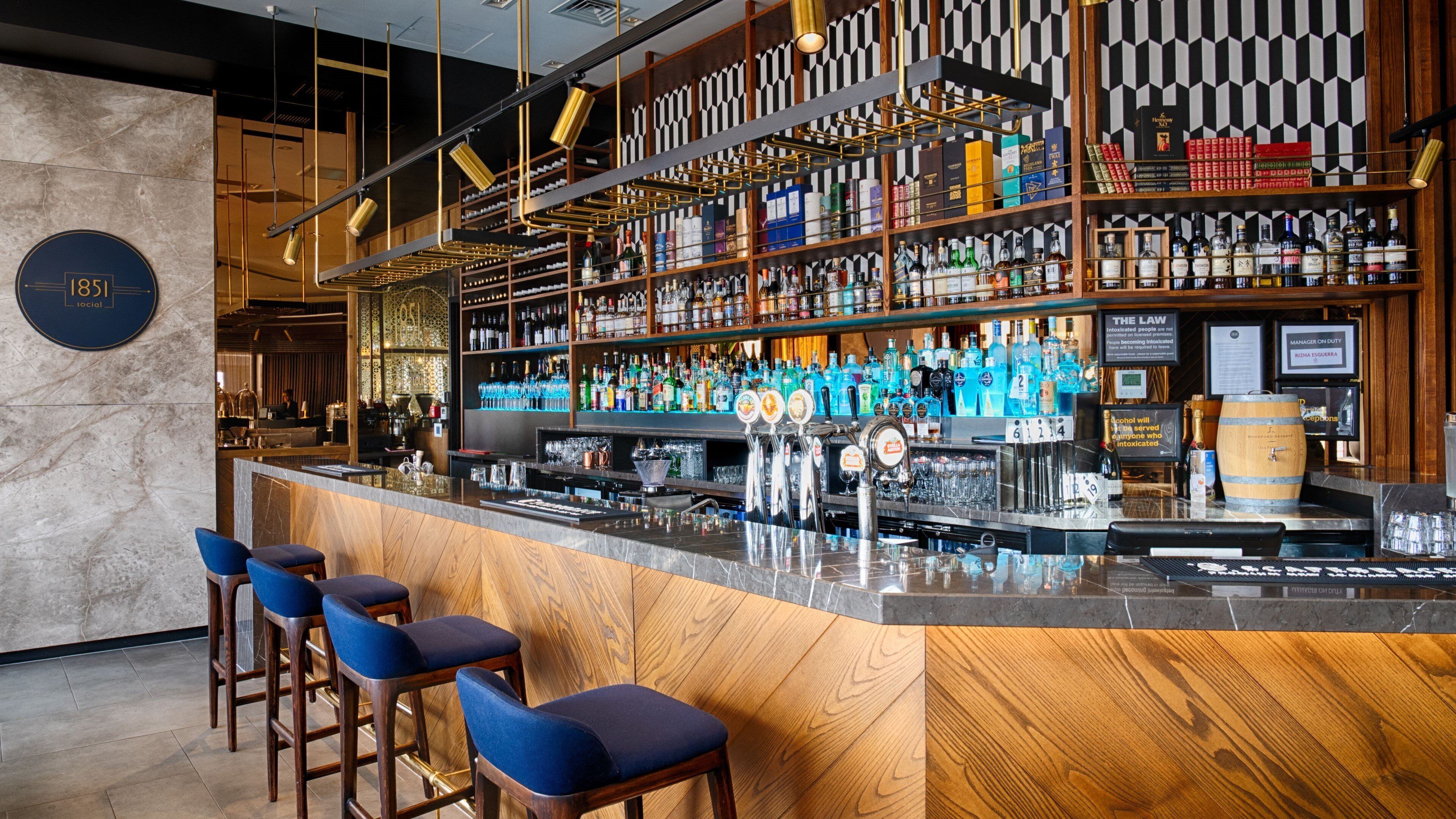 Zona de bar con pared de mármol, mostrador de madera y taburetes azules. Estantes llenos de diferentes botellas de licores.