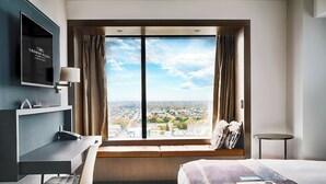 A hotel room with a large window, a desk, a bed, and a television.
