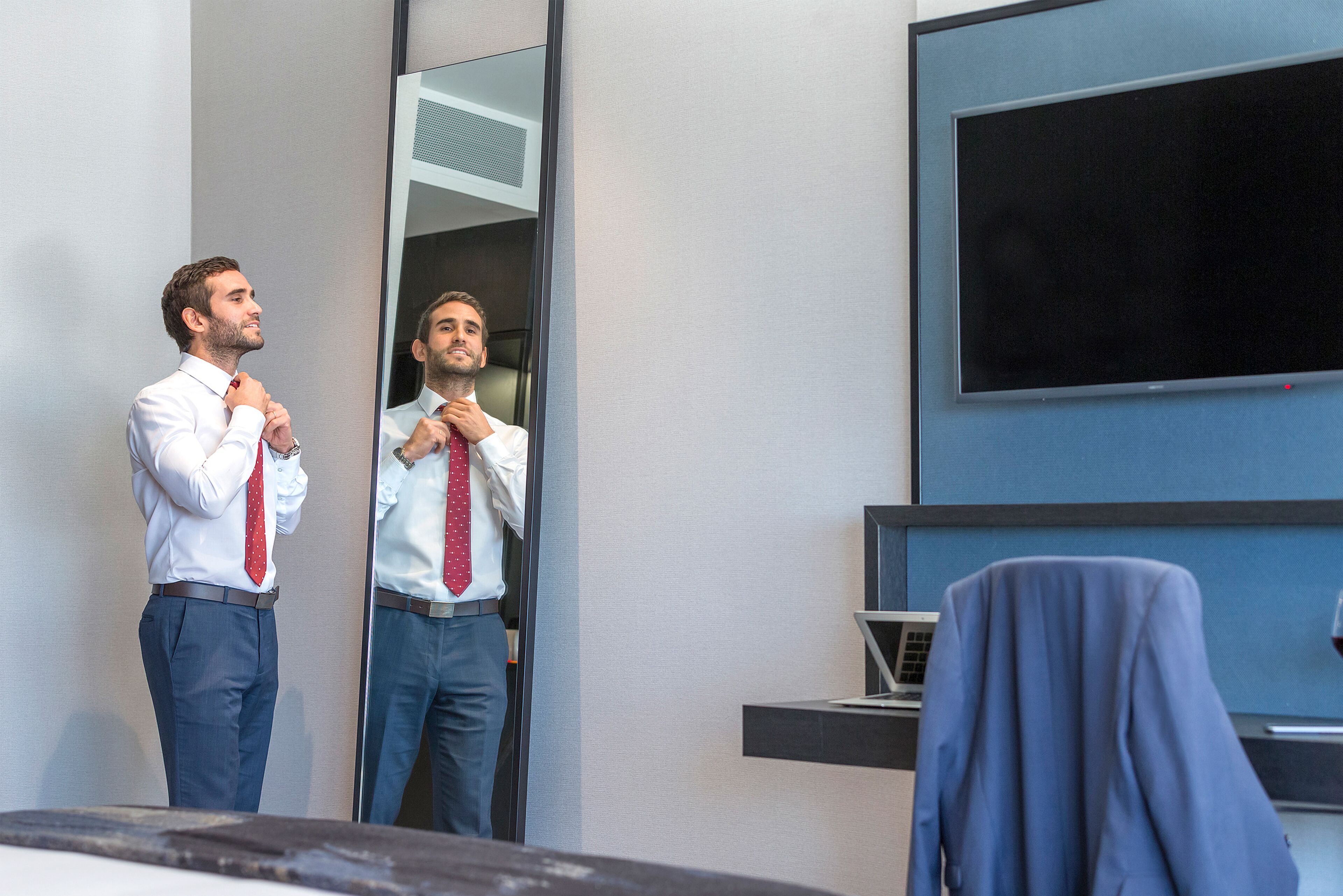A man adjusting his tie in front of a mirror in a modern office setting with a large flat-screen TV and a laptop on a desk.