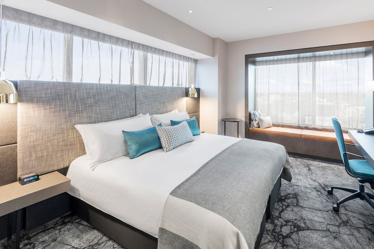 A modern hotel room with a large bed, a desk, a chair, a TV, and a window with a view.