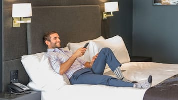 Un hombre recostado en una cama dentro de una habitación de hotel, sosteniendo un control remoto.