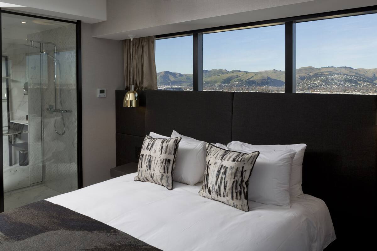 A modern hotel room with a large bed, two decorative pillows, a glass shower enclosure, a view of the city and mountains through large windows, and a dark headboard.