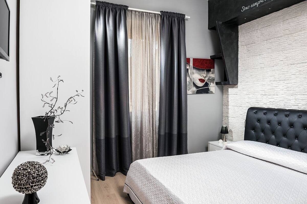 Une chambre moderne avec une tête de lit capitonnée, une table de chevet avec une lampe et une vue sur le paysage urbain à travers des rideaux transparents.
