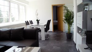 A modern living room with a sofa, a coffee table, and a dining area with chairs and a table. There is a potted plant and a TV stand. - La Domus Premium (Lotte)