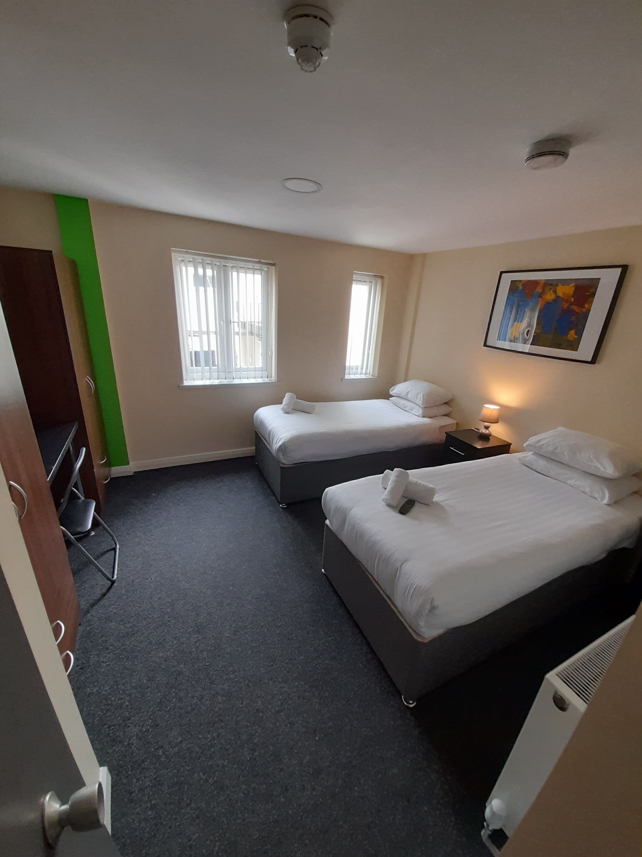 Une chambre d’hôtel avec deux lits, un bureau, une chaise, un radiateur et un tableau au mur.