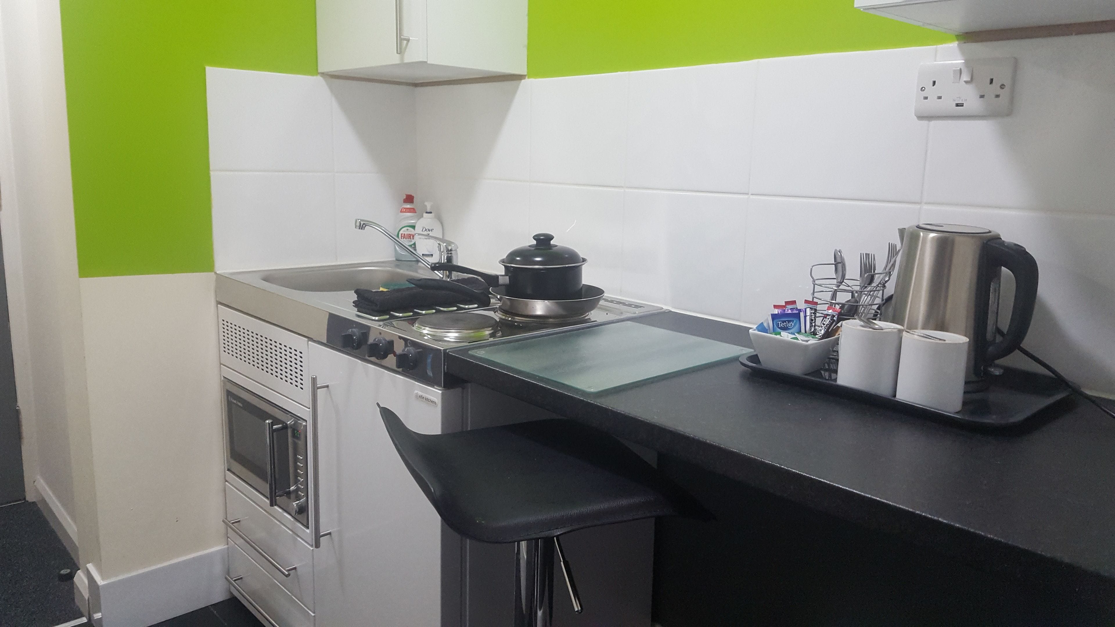 A compact kitchenette with a stove, sink, and a small oven. There are bar stools and a kettle on the counter.