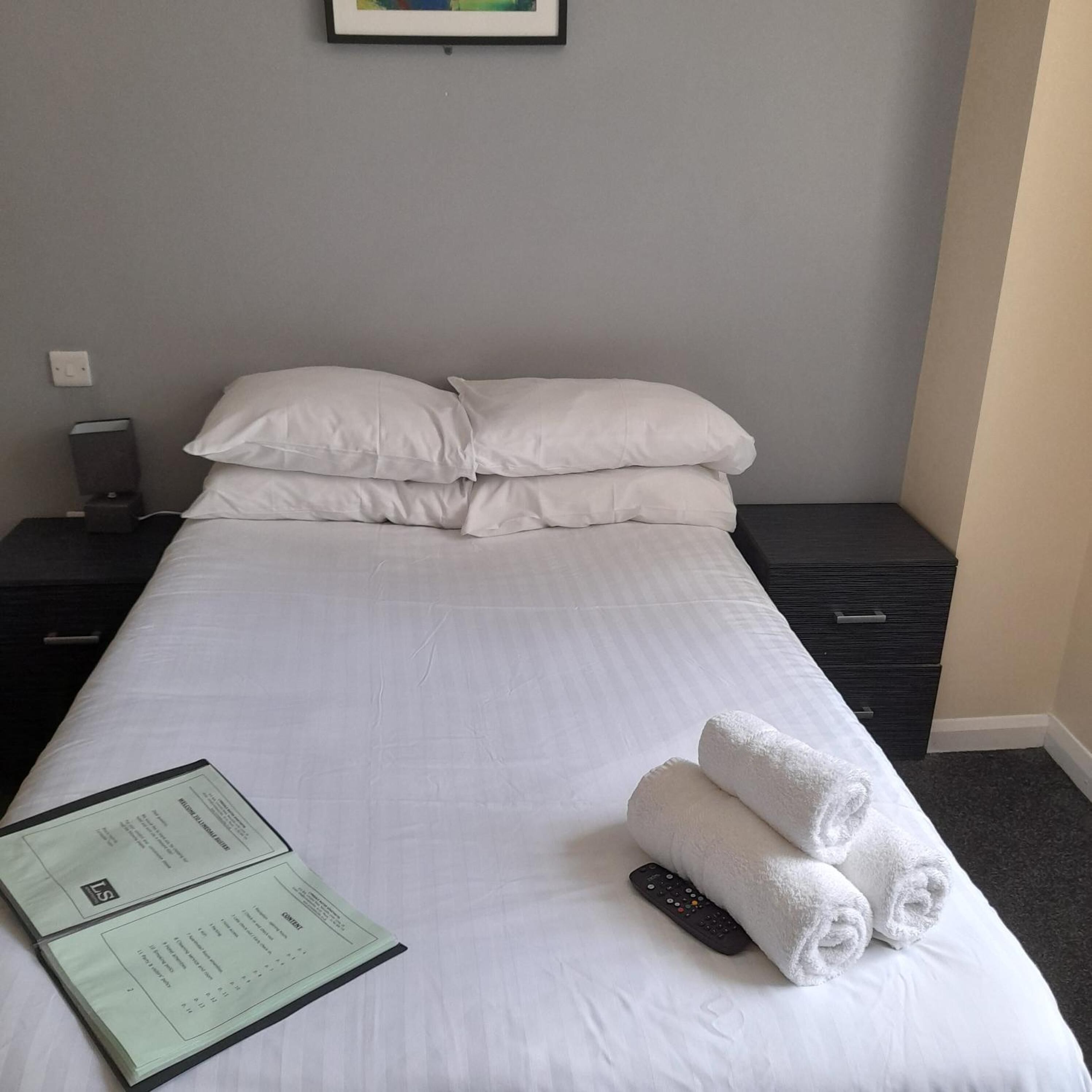 A neatly made bed with white linens, a remote control, and rolled towels on the bed.